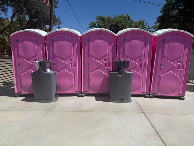 Row of four pink porta potties with two gray hand-wash stations set up on concrete for an event
